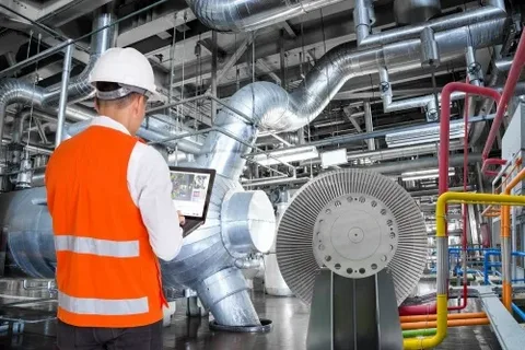Engineer in an orange vest and hard hat inspecting machinery and systems on a tablet in an industrial environment.