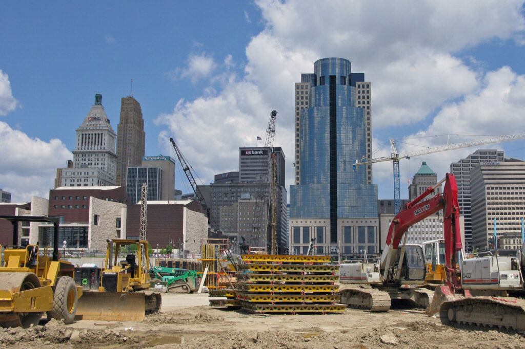 Construction site with heavy machinery including bulldozers and excavators in front of modern high-rise buildings and cranes in a downtown urban area under a partly cloudy sky