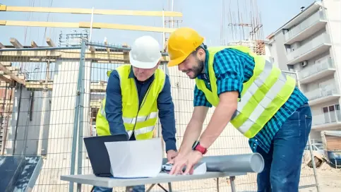 Two construction professionals reviewing blueprints and project details on a laptop at a construction site. The image highlights the collaboration and precision in providing Structural Trade Estimating Services for building projects.