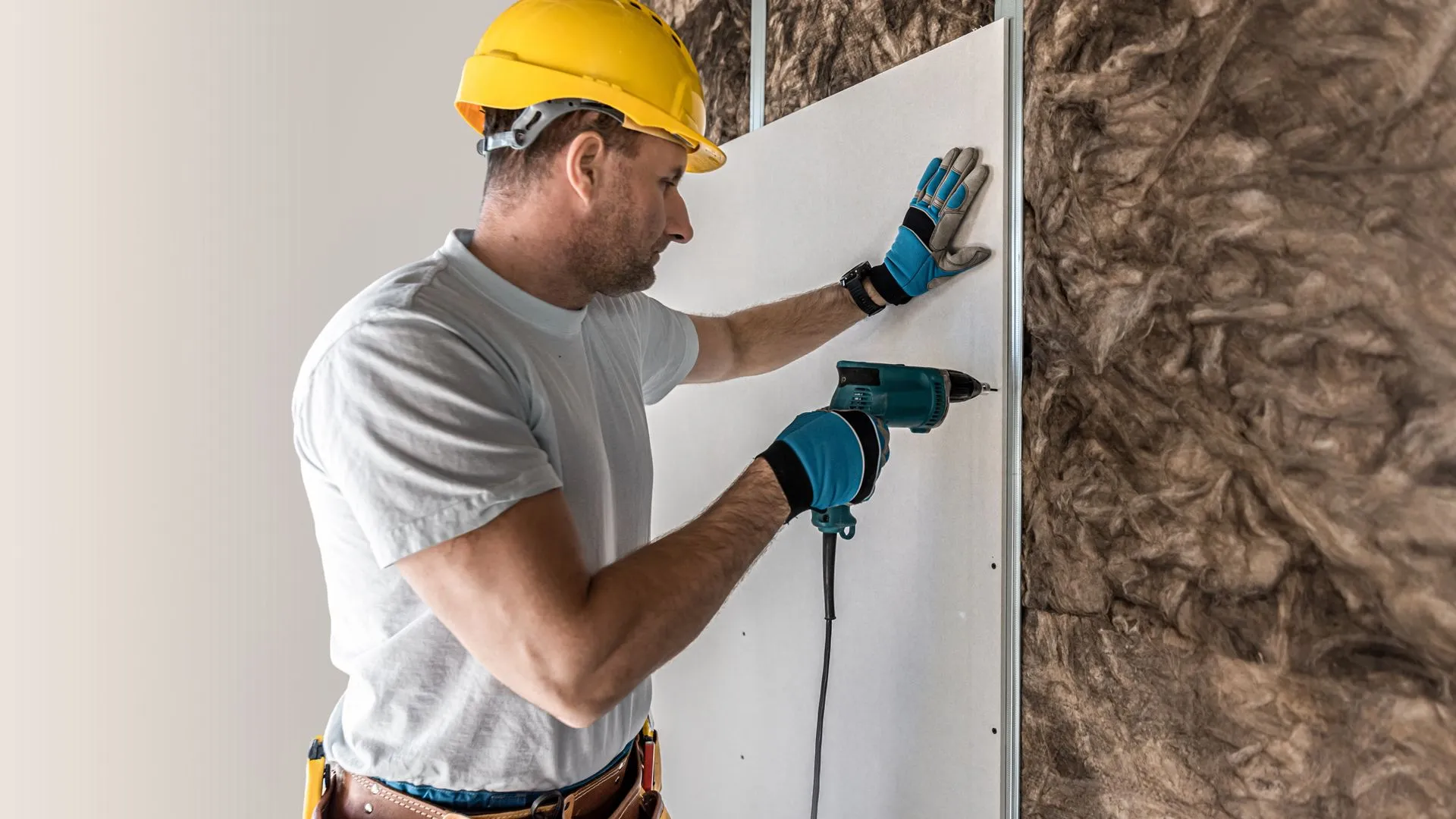 Worker in a yellow hard hat using a drill to install drywall, showcasing the process of drywall estimation for construction projects.