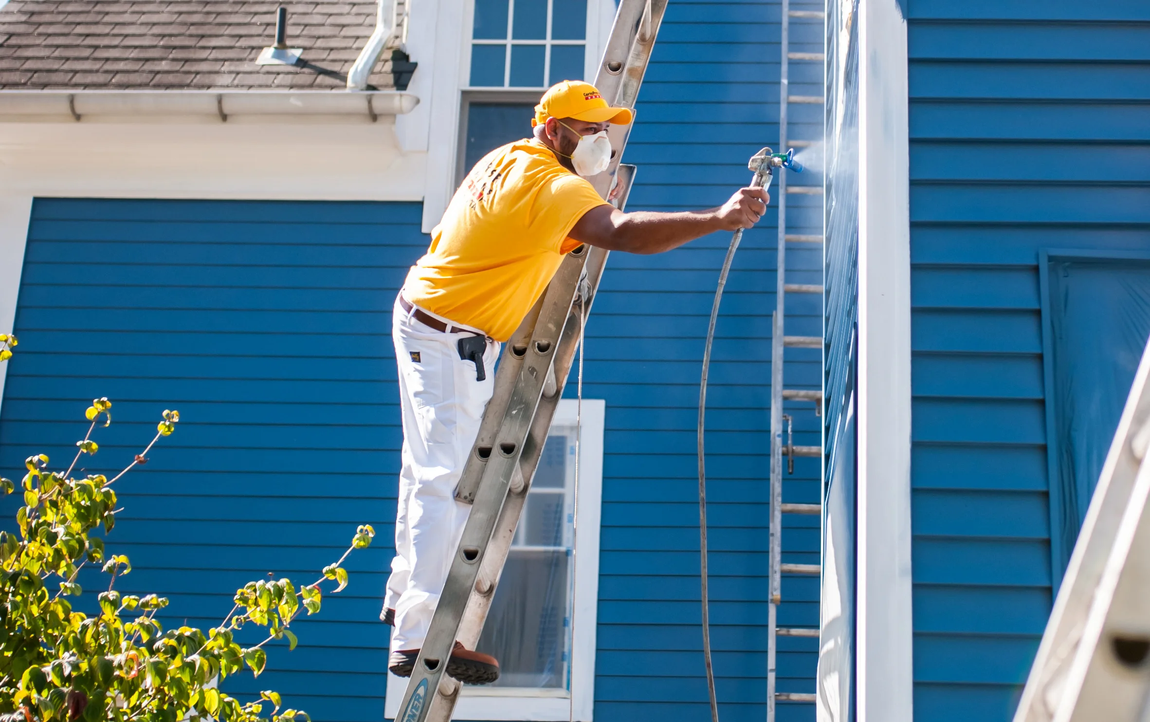 A worker in a yellow shirt and cap is painting the exterior of a building using a spray gun while standing on a ladder, wearing a face mask for protection.