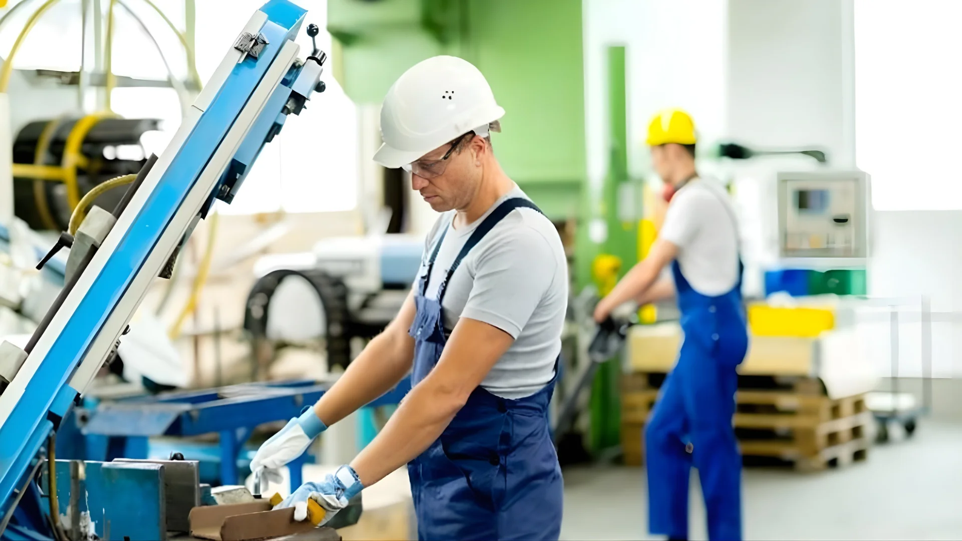 A worker in a hard hat and safety gear is operating machinery in a factory, while another worker is in the background, indicating an industrial setting focused on manufacturing processes.