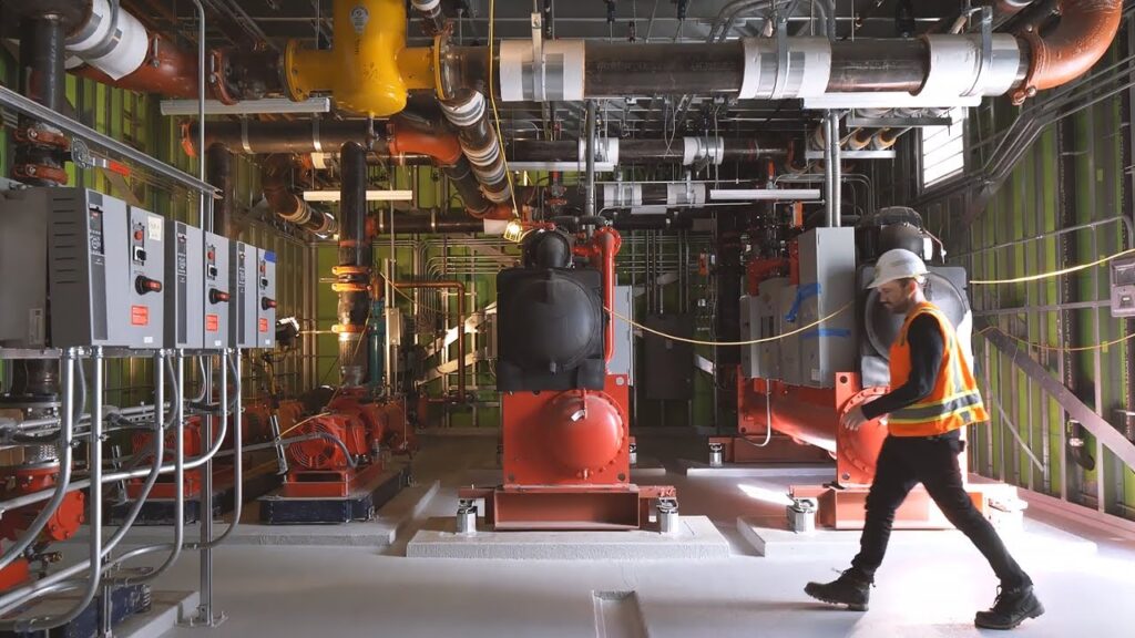 A worker walking through a mechanical room with various mechanical, electrical, and plumbing (MEP) systems in place, showcasing the importance of MEP Estimation for accurate construction planning by EZ Estimation.
